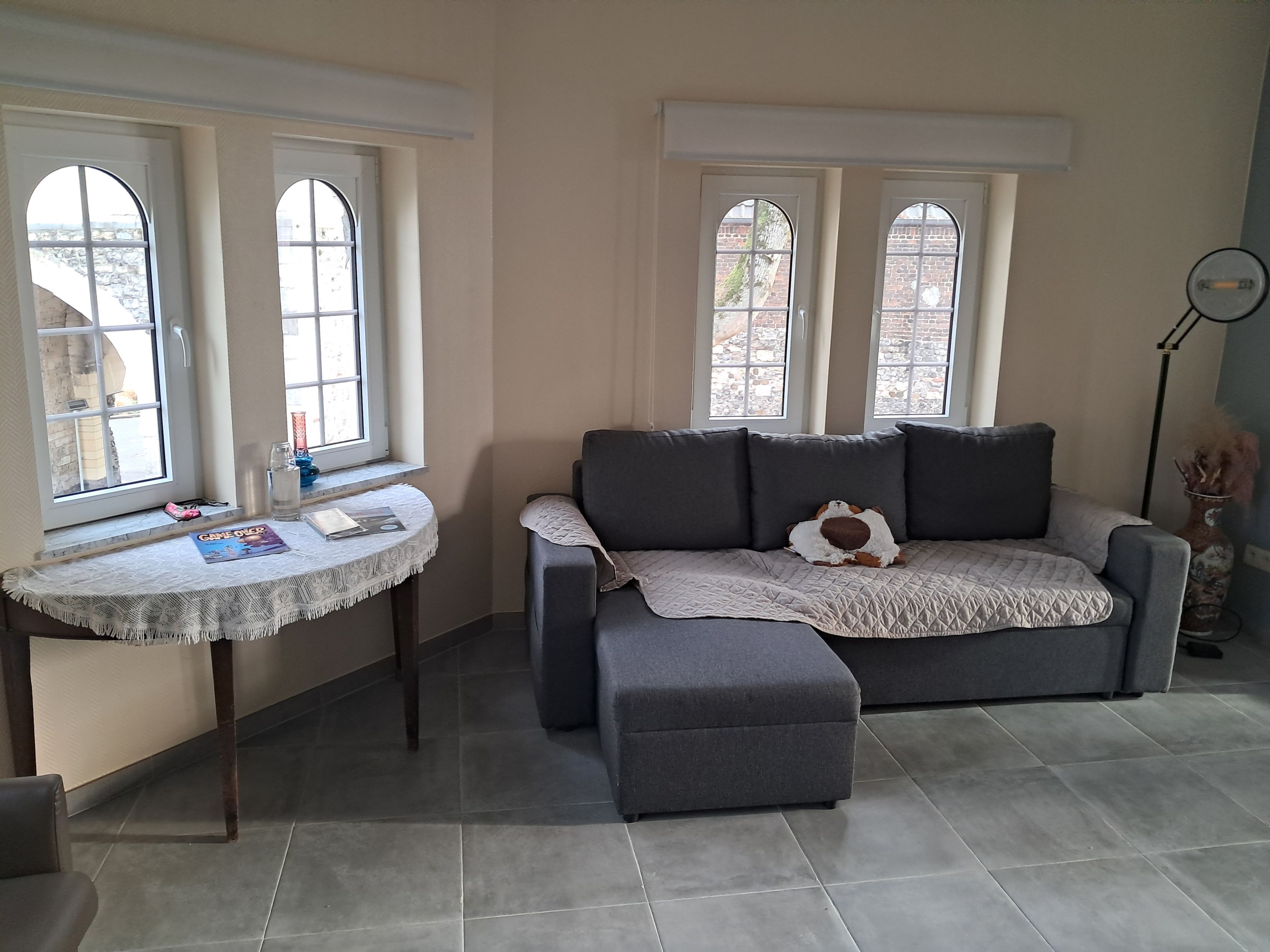 Una habitación con un sofá gris, una mesita redonda con mantel de encaje y una ventana con vista a un muro de ladrillo.
