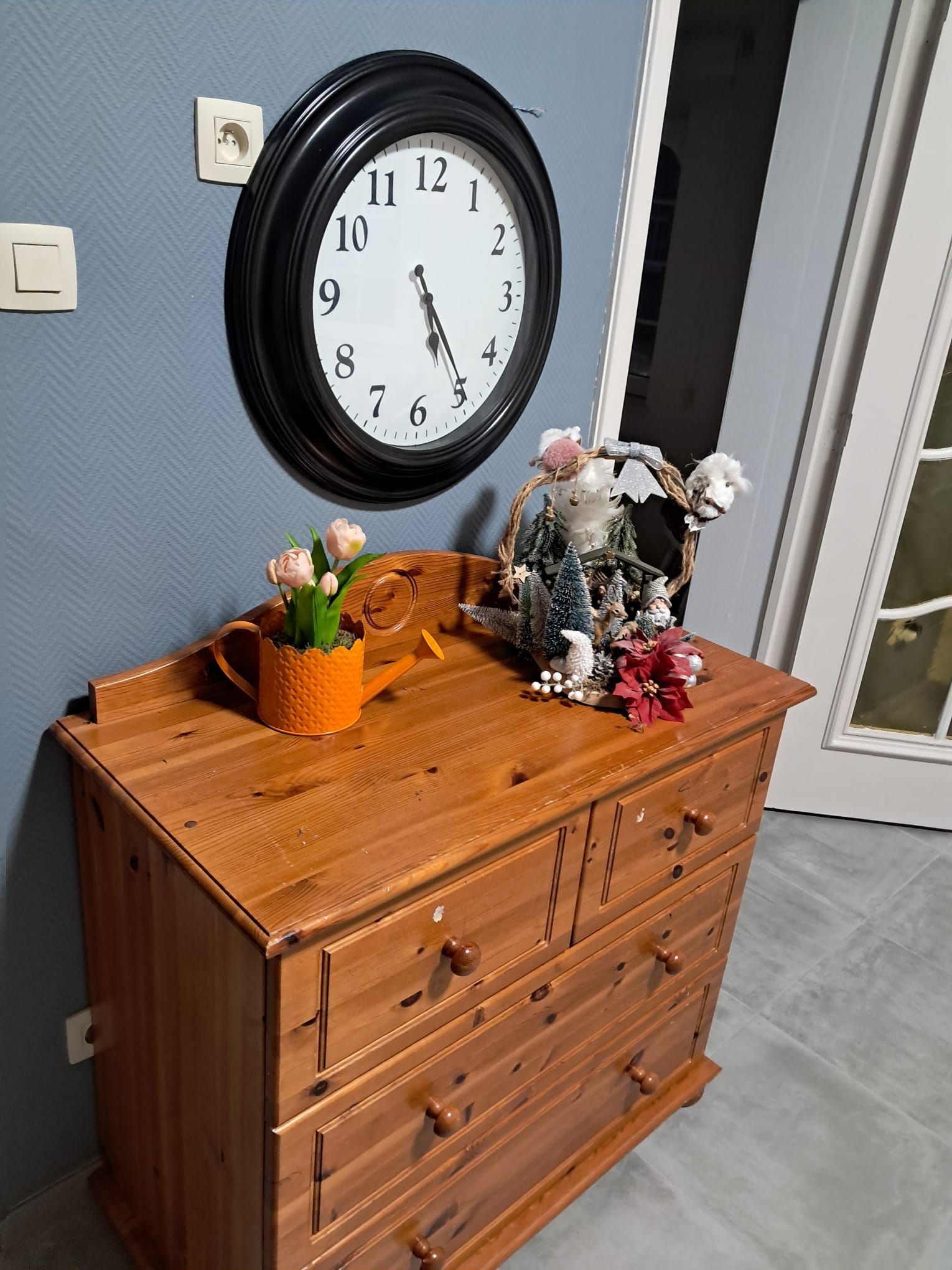 Ein hölzerner Kommode mit Schubladen, eine Wanduhr und eine kleine Dekoration mit Blumen und einer Miniatur-Weihnachtslandschaft.