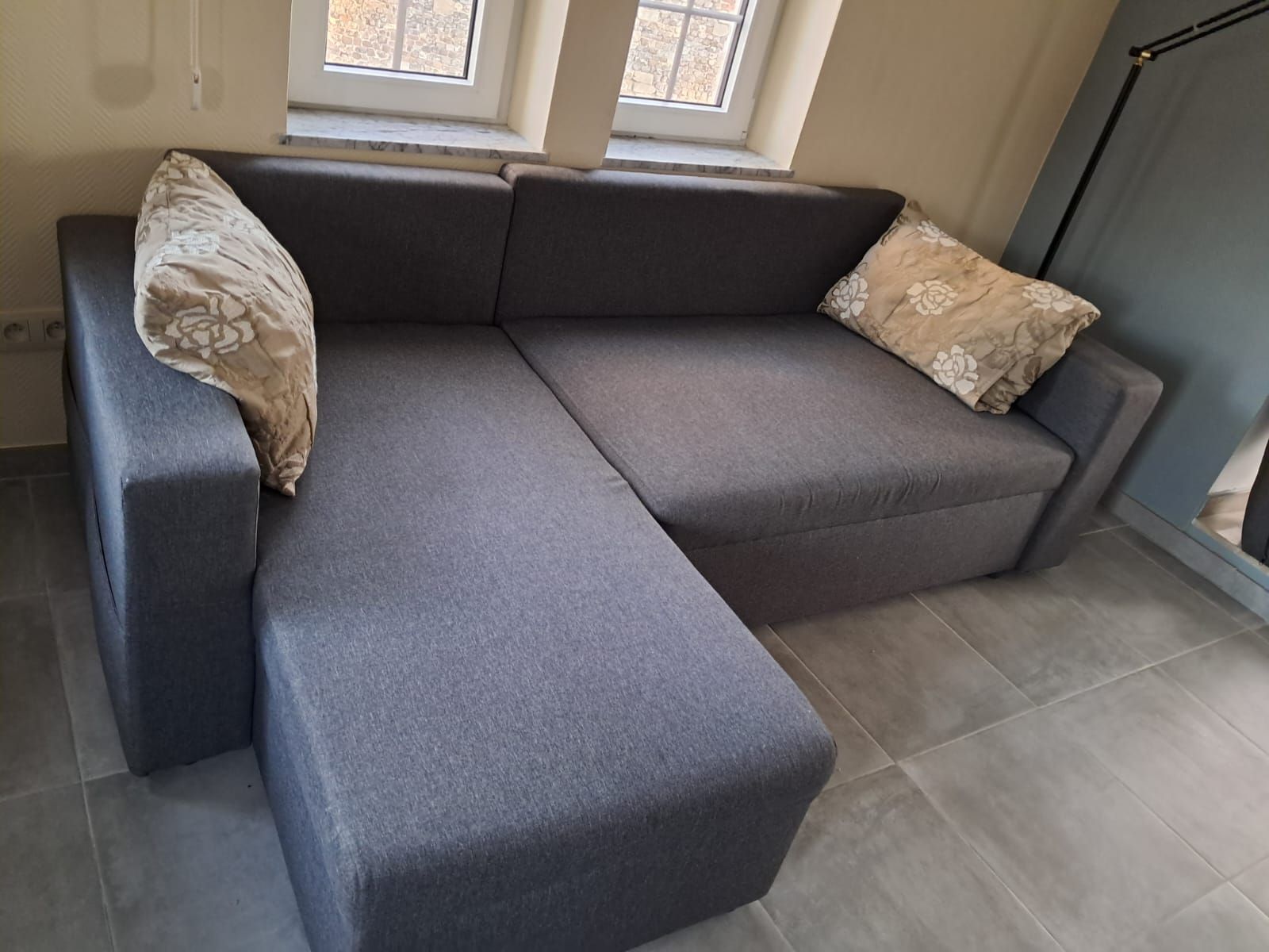 Un sofá seccional gris con cojines estampados en una habitación con piso de baldosas y dos ventanas.