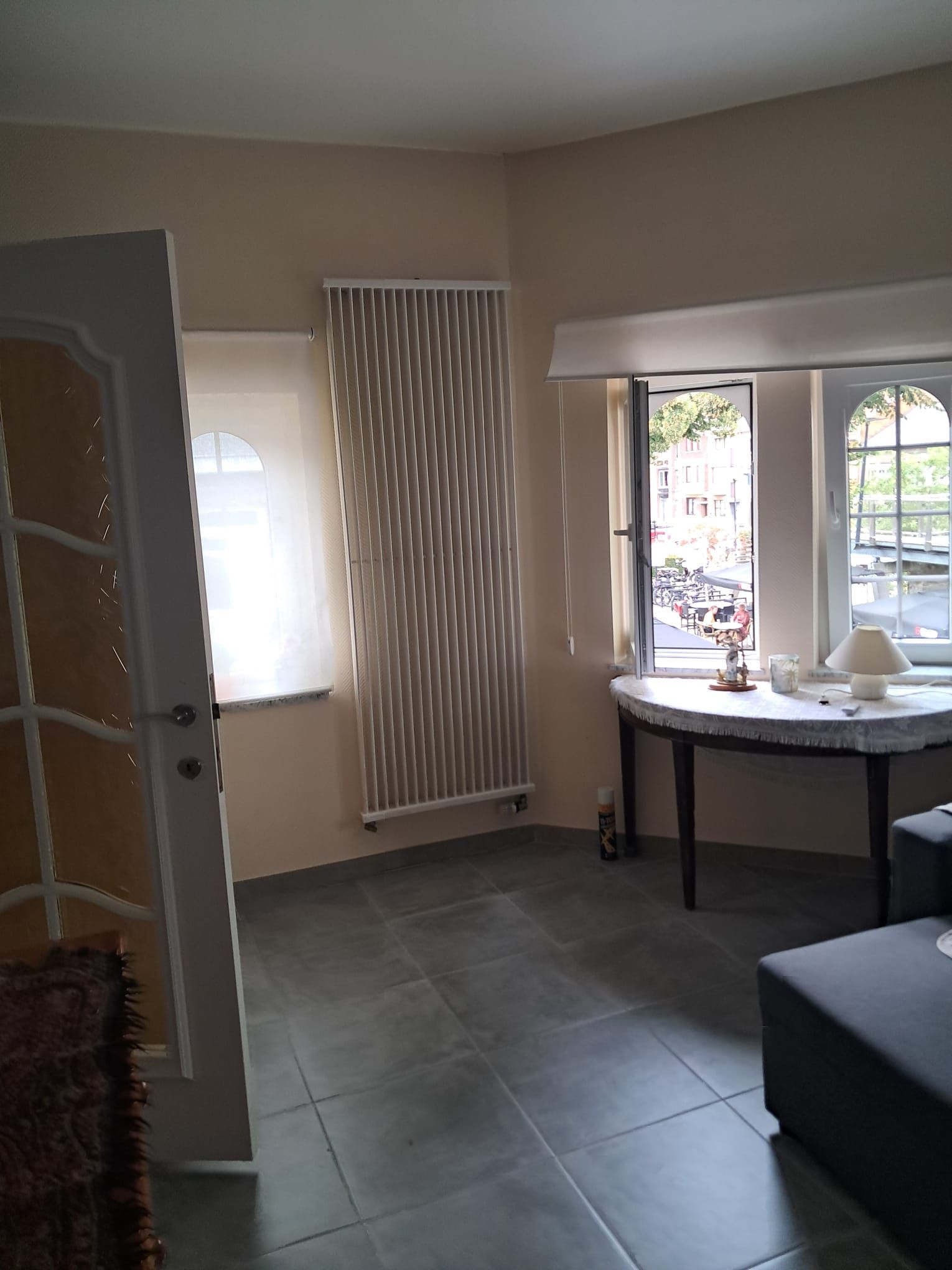 Una habitación con un radiador, un ventanal con vista a la calle, y una pequeña mesa con una lámpara y una taza.