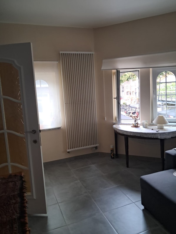 Una habitación con un radiador, un ventanal con vista a la calle, y una pequeña mesa con una lámpara y una taza.