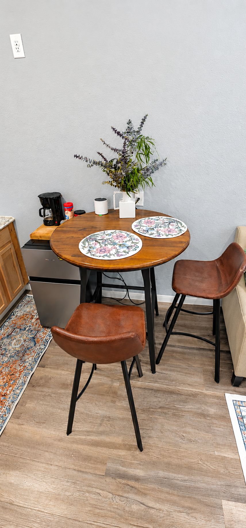 Un coin repas avec une table en bois, deux chaises, une machine à café, une plante en pot et un tapis à motifs.