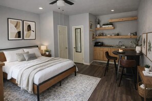 A bedroom with a bed, bedside tables, a nightstand, a desk with a chair, a small table with a candle, and a kitchen area with shelves and a sink.