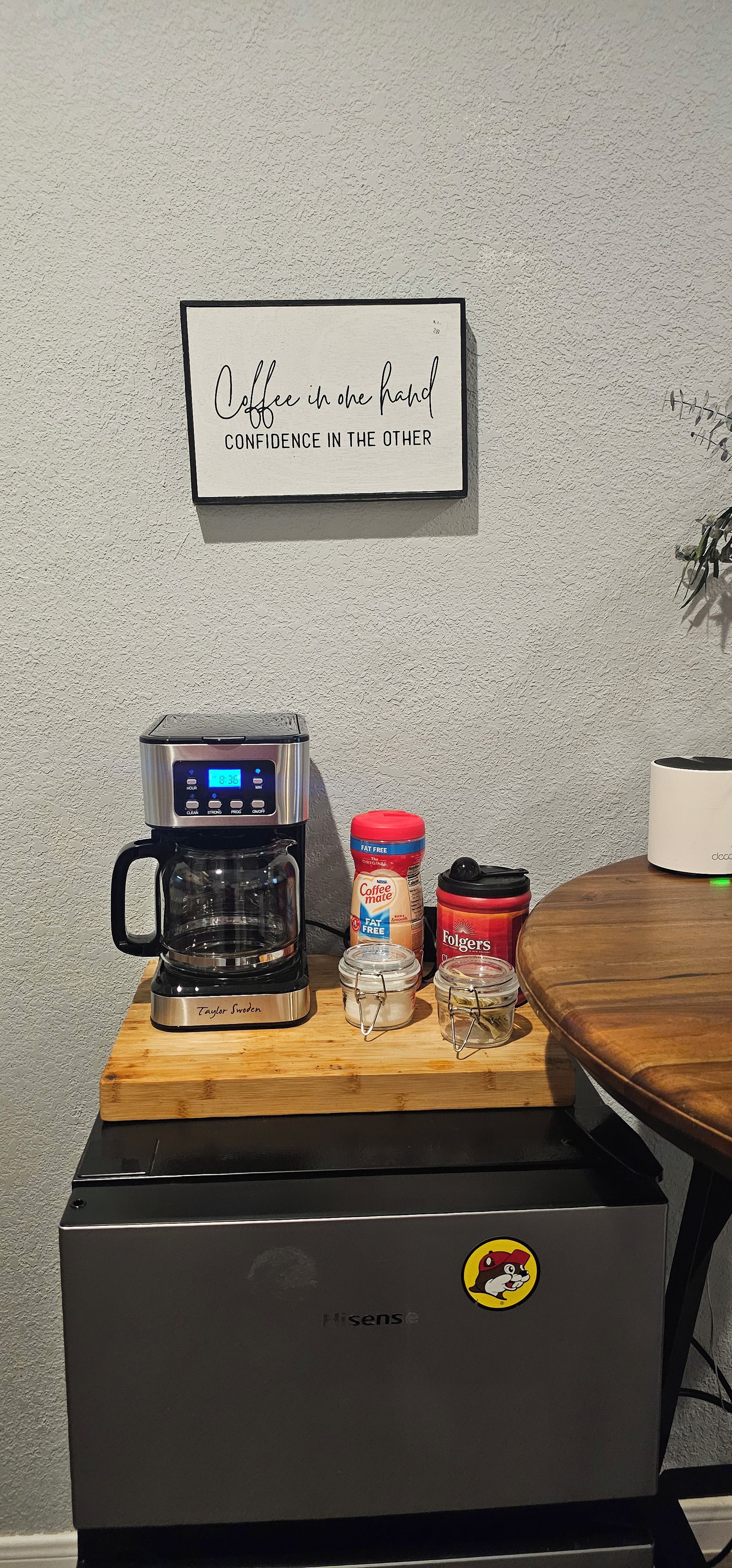 Eine Kaffeemaschine, ein Behälter mit Kaffeeweißer, ein Zuckerbehälter und eine Kaffeemühle stehen auf einem Holztisch. Ein Schild darüber verkündet: 'Kaffee in der einen Hand, Zuversicht in der anderen.'