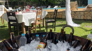 Uma área de refeições ao ar livre com mesas e cadeiras, uma piscina e um bar com garrafas em gelo.