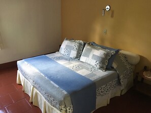 Un llit amb un llençol i coixins estampats en blau i blanc, un rellotge de paret i una taula petita amb un objecte decoratiu.