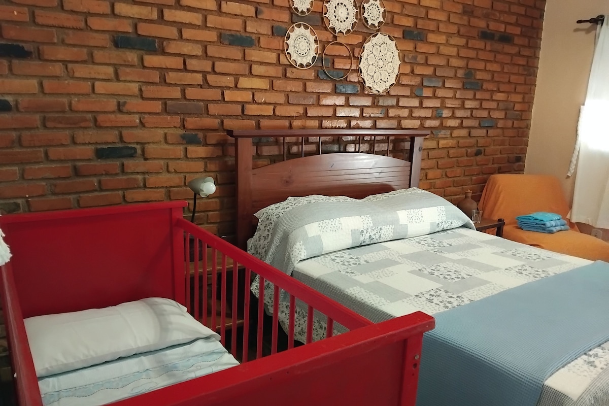 A room with a brick wall, a wooden bed with a headboard, a red crib, and a chair with a cushion.