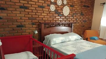 A room with a brick wall, a wooden bed with a headboard, a red crib, and a chair with a cushion.
