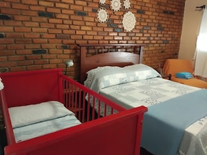 A room with a brick wall, a wooden bed with a headboard, a red crib, and a chair with a cushion. - Fazenda Hotel Jatahy  (Paraíba do Sul)