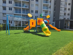 A playground with a slide and swings, surrounded by a well-maintained lawn, with a multi-story building in the background. - A beautiful new apartment in Uruka Point, San Jose. Great location! (San José)