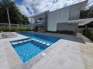 Et moderne poolområde med en klar, blå pool, et hvidt hegn og en bygning med et unikt arkitektonisk design.