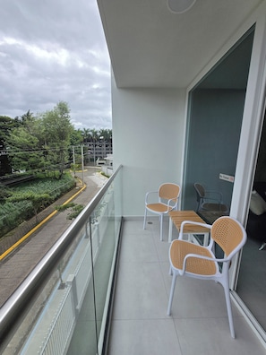Ein Balkon mit Glasgeländer, zwei Korbstühle, ein kleiner Tisch und Blick auf eine Straße und Grünflächen.