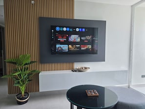A modern living room with a flat-screen TV mounted on the wall, a potted plant, a glass coffee table, and a remote control on the table. - A beautiful new apartment in Uruka Point, San Jose. Great location! (San José)