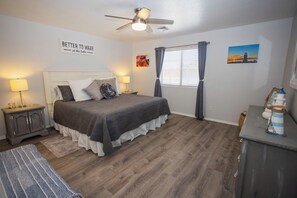 Ein Schlafzimmer mit Bett, Deckenventilator, Nachttisch, Lampe, einem Fenster mit Vorhängen und einem Wandbild.