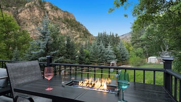 An outdoor seating area with a fire feature, a glass table, and a view of a mountainous landscape.