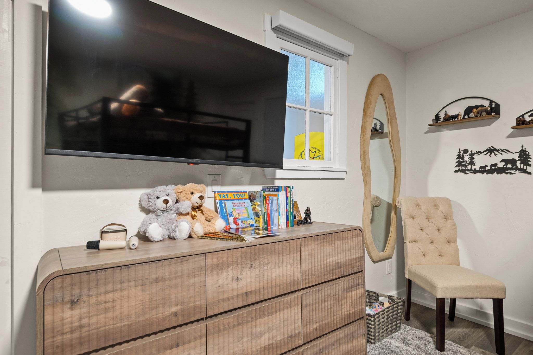 A room with a television mounted on the wall, a wooden dresser with books and stuffed animals, a chair, and a mirror.