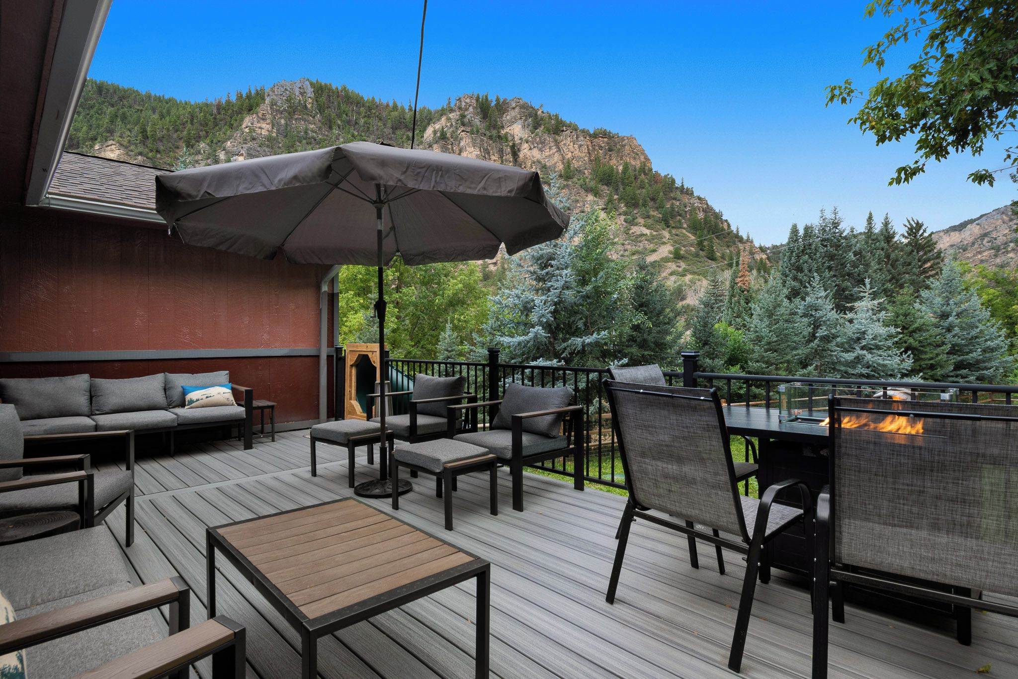 An outdoor deck with a sofa, chairs, and a table, overlooking a mountainous landscape.