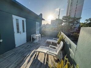 Un balcon doté d’un plancher en bois, de chaises blanches, d’une petite table et de plantes en pot.