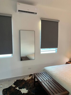 Ein Schlafzimmer mit einem Bett, einer hölzernen Bank, einem großen Spiegel und zwei Fenstern mit Jalousien.