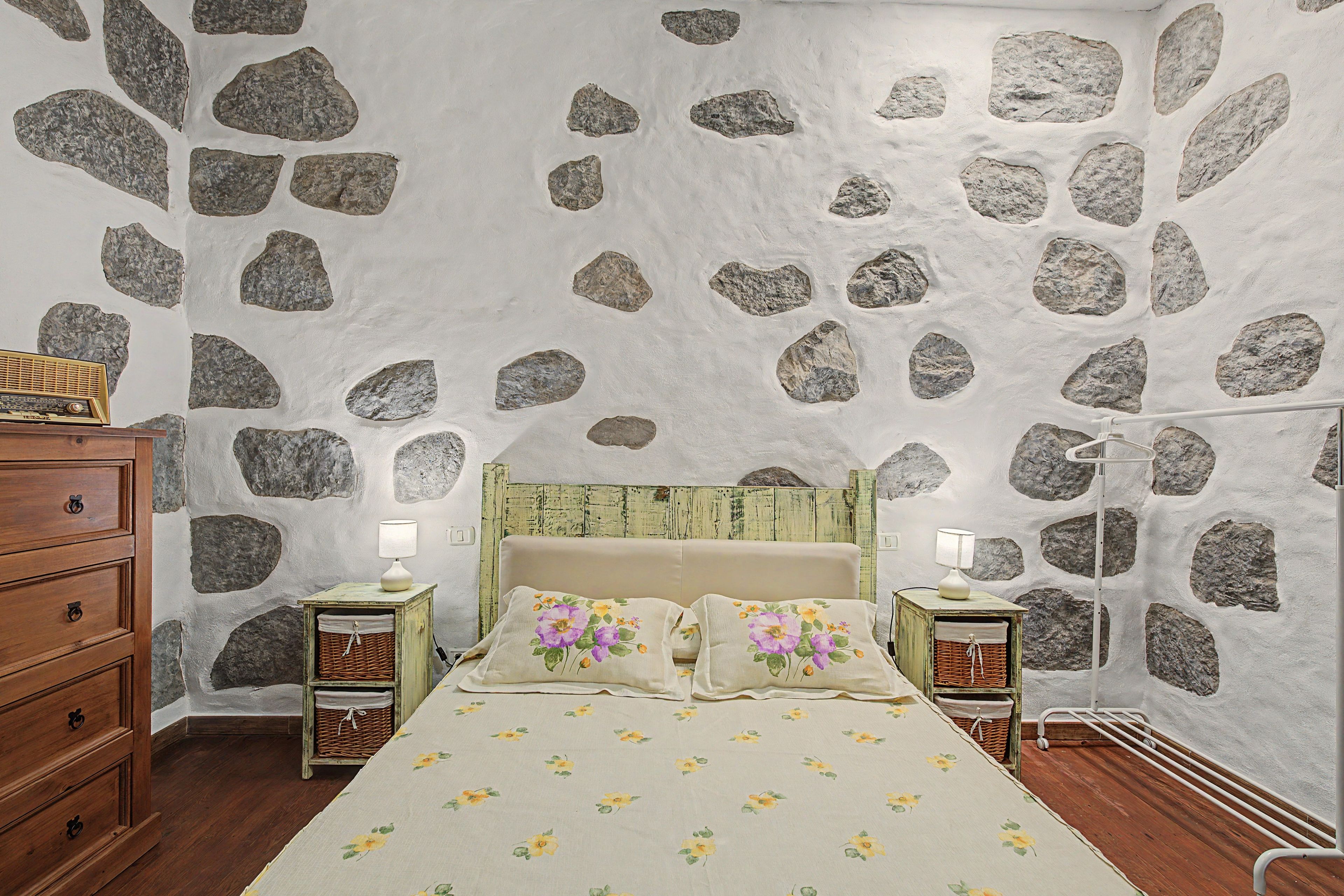 Une chambre avec un mur en pierre, un lit avec une literie à motifs floraux, des tables de chevet en bois et une commode.