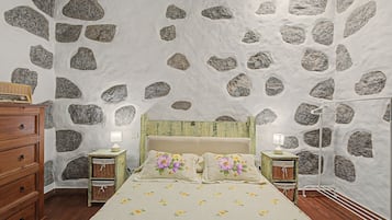 Une chambre avec un mur en pierre, un lit avec une literie à motifs floraux, des tables de chevet en bois et une commode.