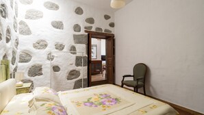 A bedroom with a stone wall, a bed with floral bedding, a chair, and a hanging light fixture. - Vacation Home 'Gloria' with Mountain Views, Shared Terrace & Wi-Fi (Arucas)