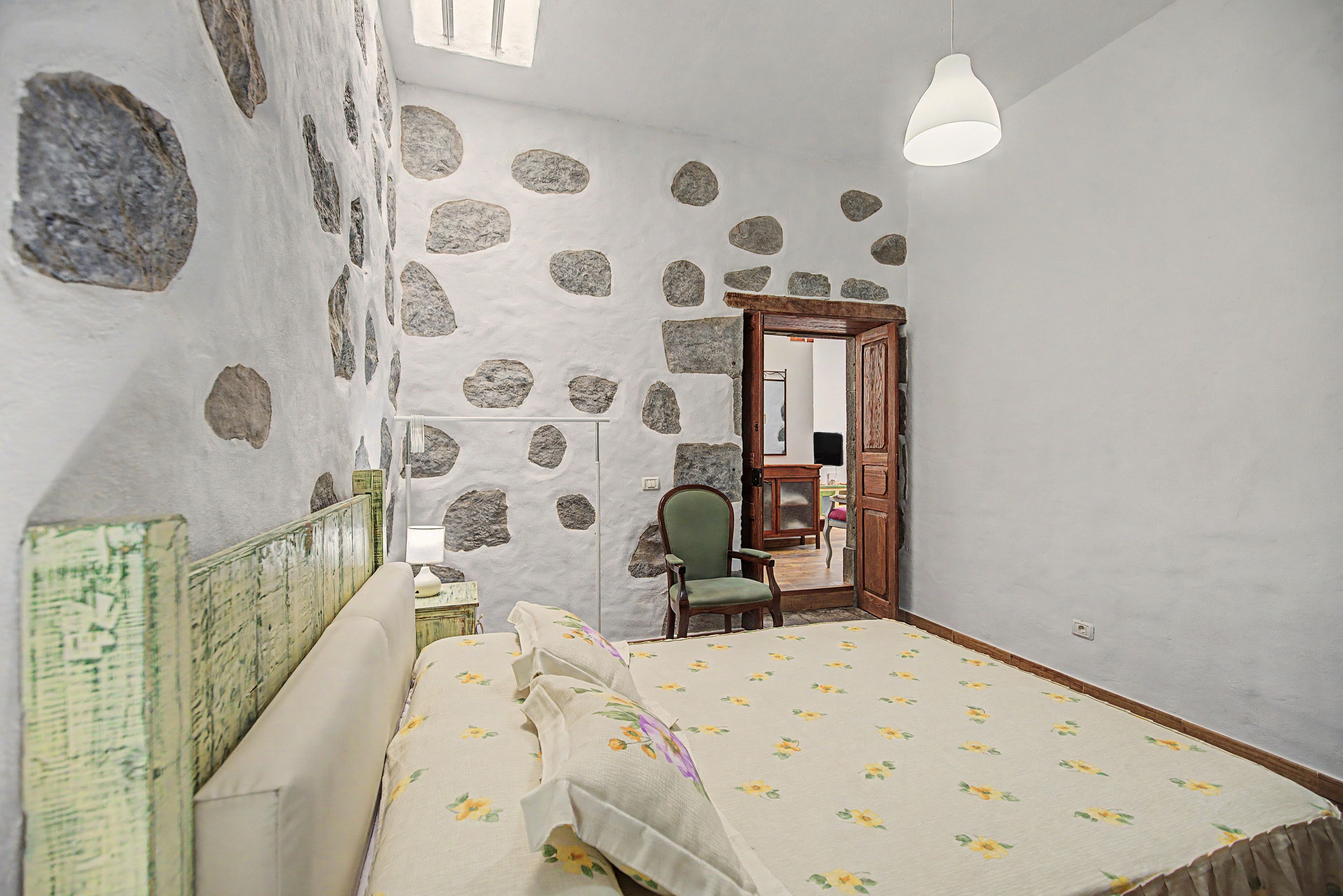 Una habitación con una cama, una silla, una puerta de madera y una pared de piedra.