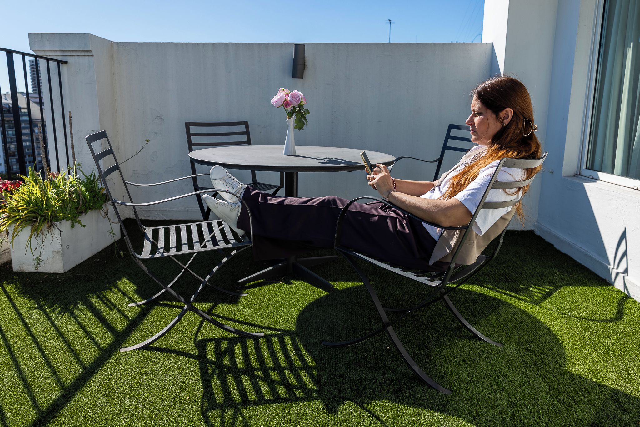 A person seated at a round table with a chair, on a balcony with artificial grass, a white wall, and a window.