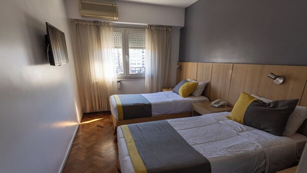 A hotel room with two beds, a wooden headboard, a window with curtains, a wall-mounted TV, and a phone on the nightstand. - AURA HOTEL RECOLETA (Buenos Aires)