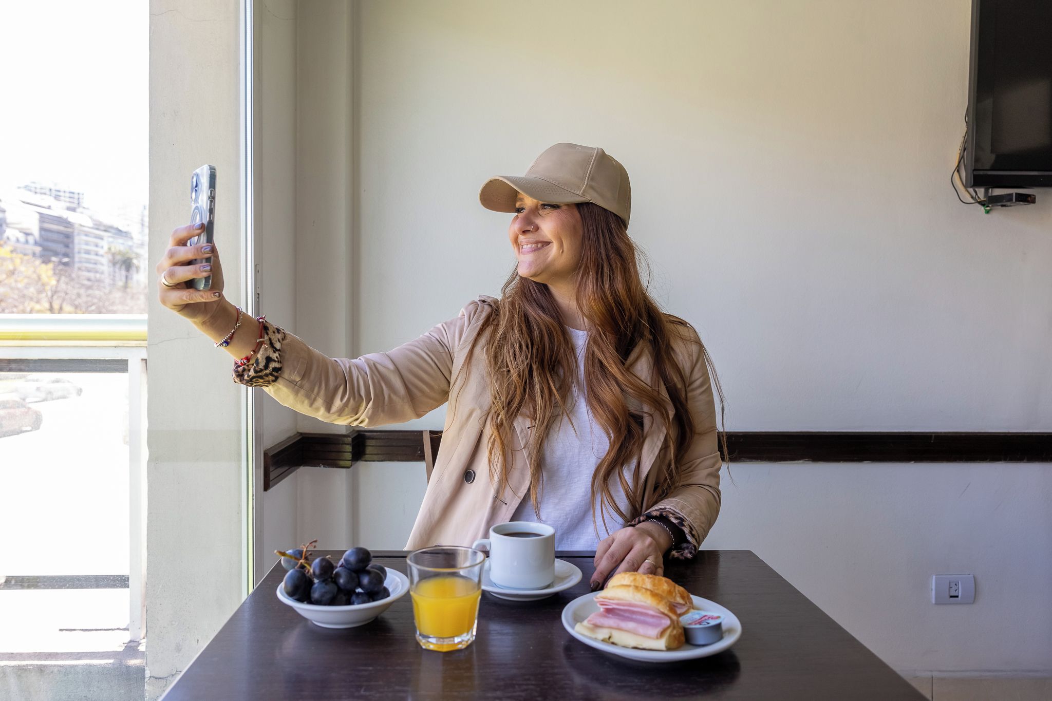 A person taking a selfie at a table with breakfast items including grapes, orange juice, coffee, and a sandwich.