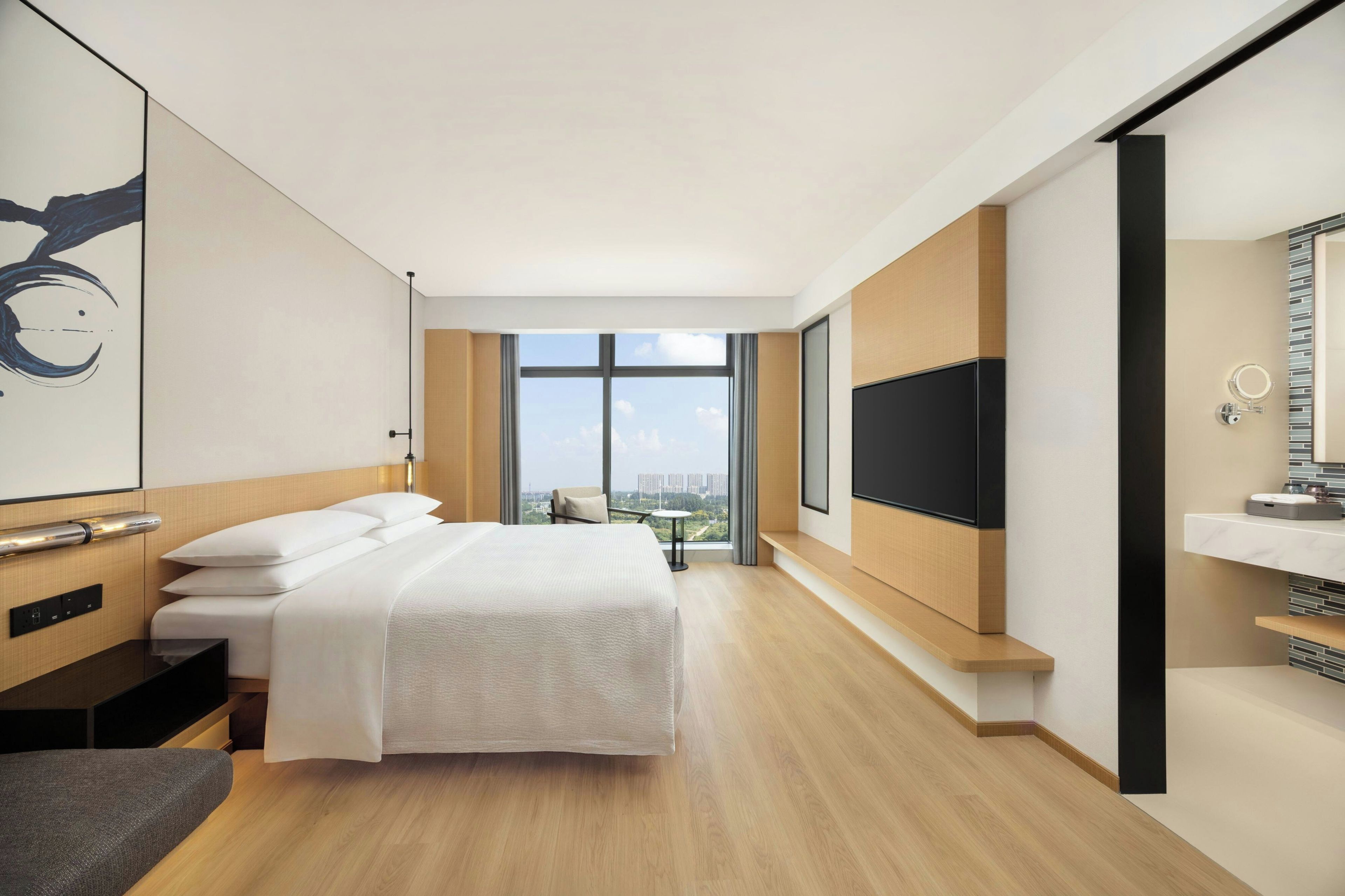 A hotel room with a large bed, a TV, a bathroom with a mirror, and a view of the city.