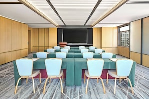 A conference room with a large table, chairs, a screen, and a view of buildings through the windows.
