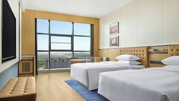 A hotel room with two beds, a bench, a large window with a city view, and a flat-screen TV.