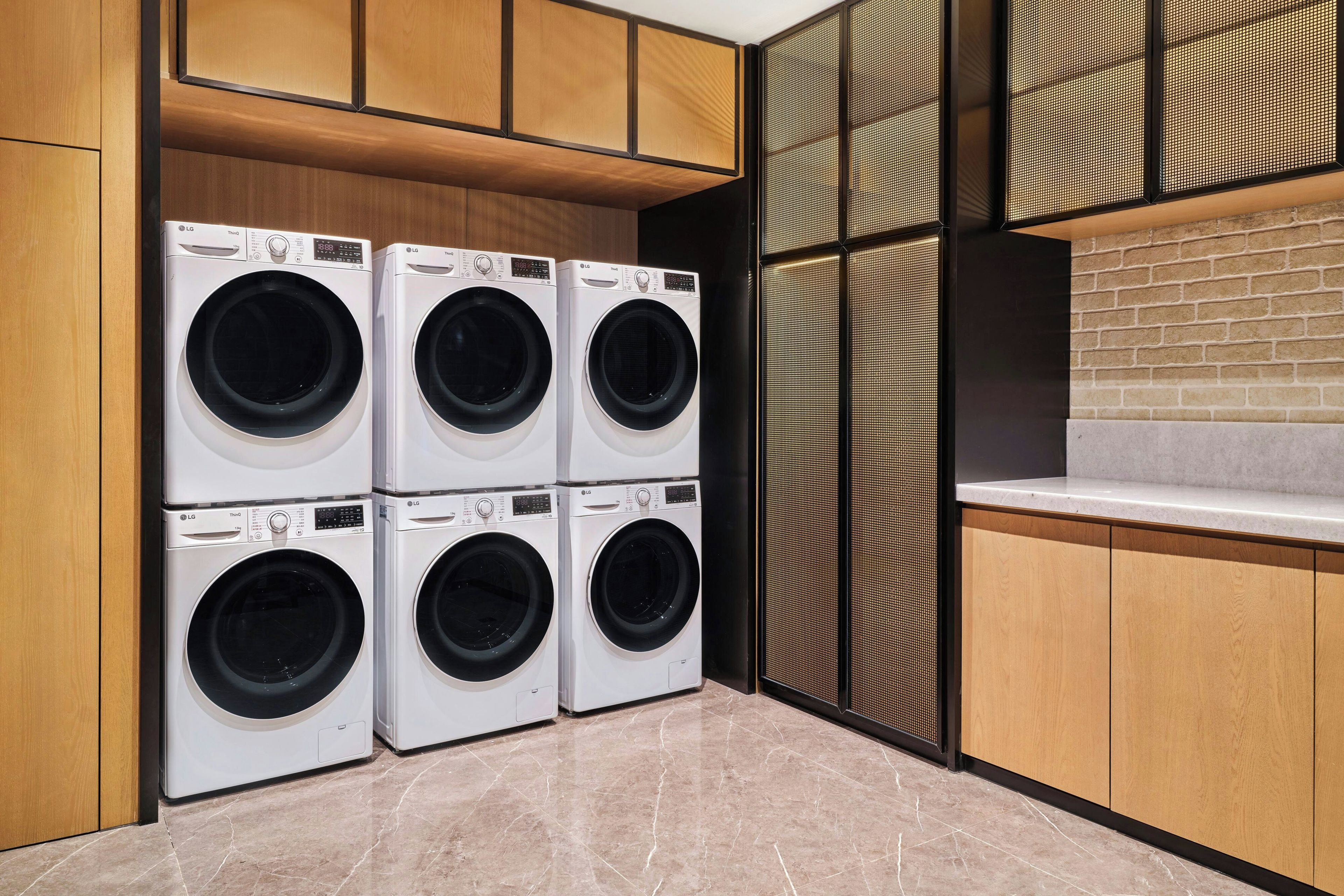 Un cuarto de lavandería con cuatro lavadoras LG ThinQ, armarios de madera, piso de baldosas y pared de ladrillo.