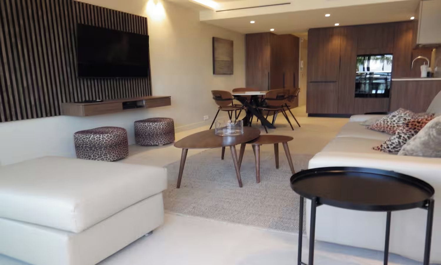 A modern living room with a sofa, a coffee table, a TV mounted on the wall, and a dining area with chairs and a table.