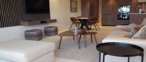 A modern living room with a sofa, a coffee table, a TV mounted on the wall, and a dining area with chairs and a table.