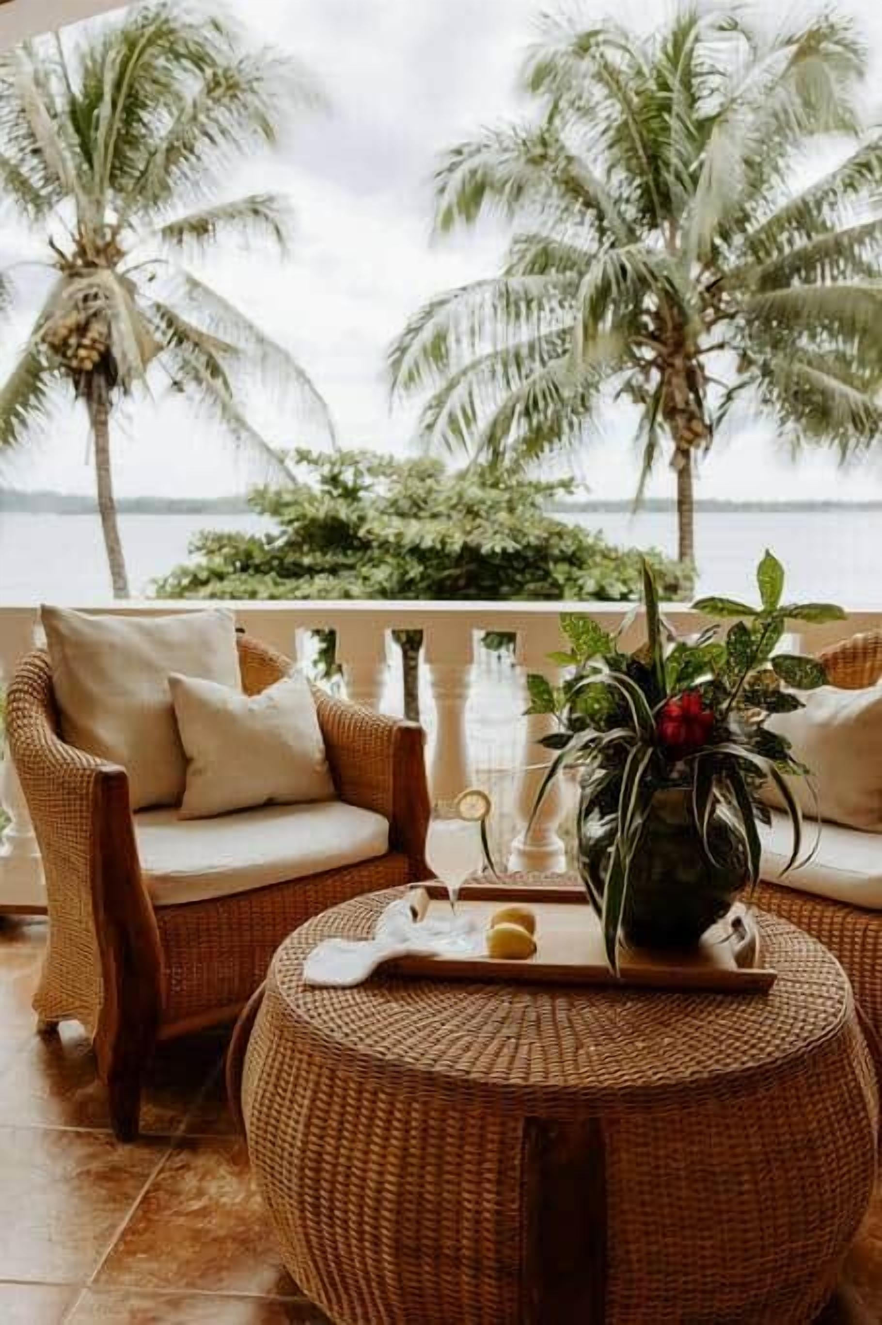 A wicker sofa with cushions, a round wicker table with a vase and a tray, and a view of palm trees and water.