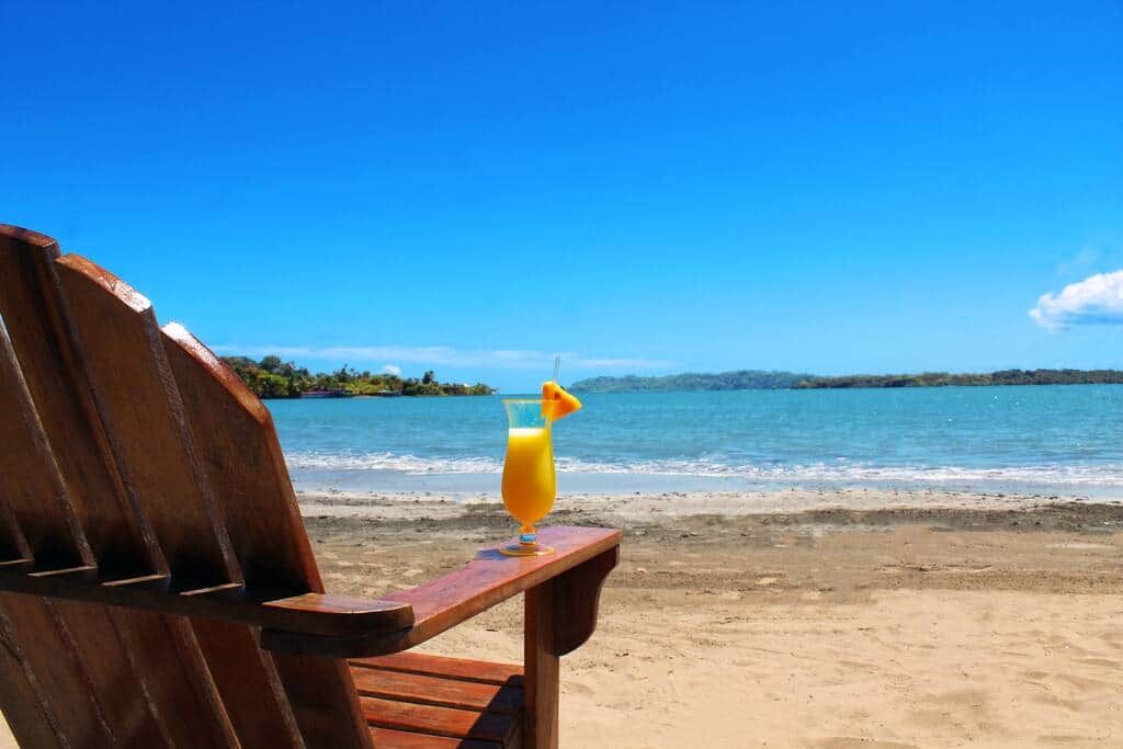 A wooden chair with a glass of orange juice on a beach.