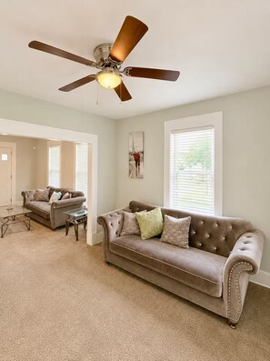 A living room with a brown leather sofa, a glass coffee table, a ceiling fan, and large windows.