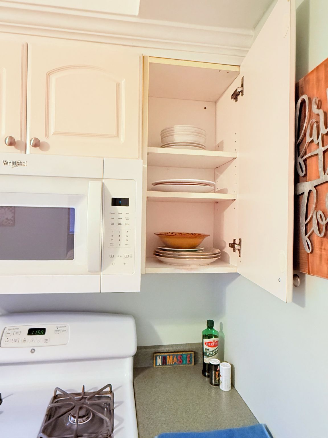 Una cocina con gabinetes blancos, un microondas, una estufa y una repisa con platos y un tazón.