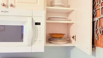 Una cocina con gabinetes blancos, un microondas, una estufa y una repisa con platos y un tazón.