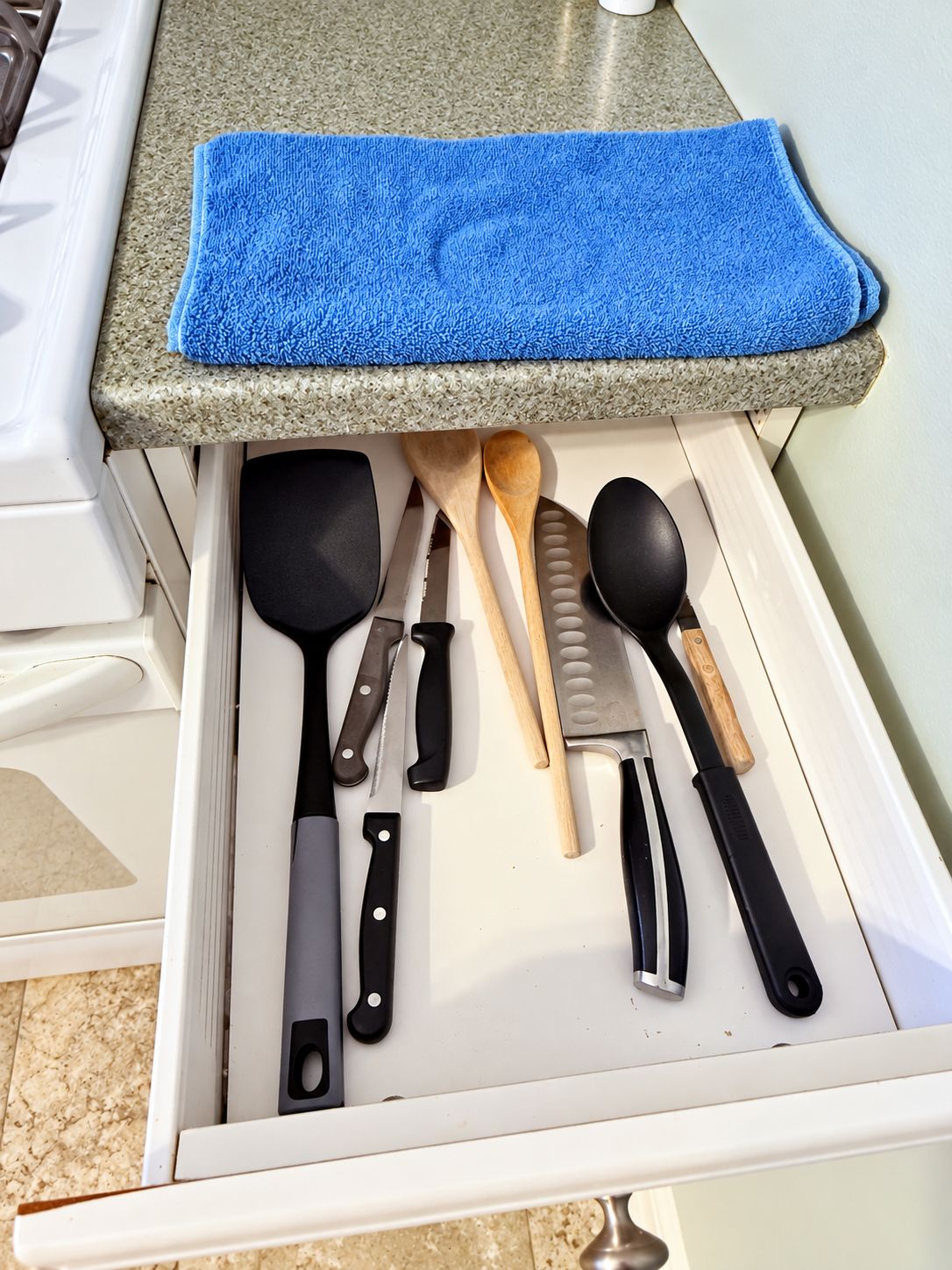 Un cajón de cocina con varios utensilios, incluyendo una toalla azul, una espátula, un cuchillo, una cuchara de madera y una cuchara para servir.