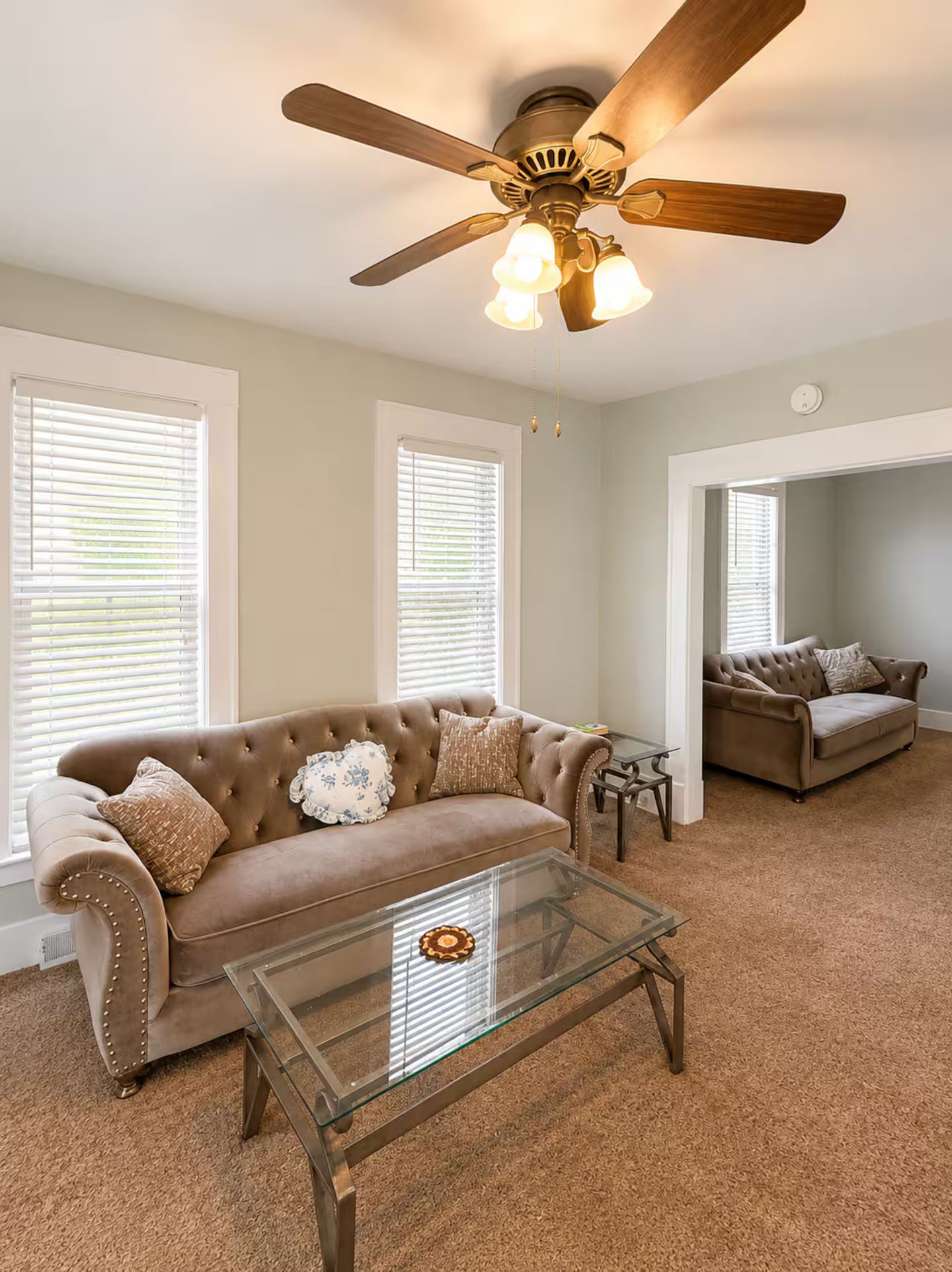 A living room with a brown sofa, a glass coffee table, and a ceiling fan.