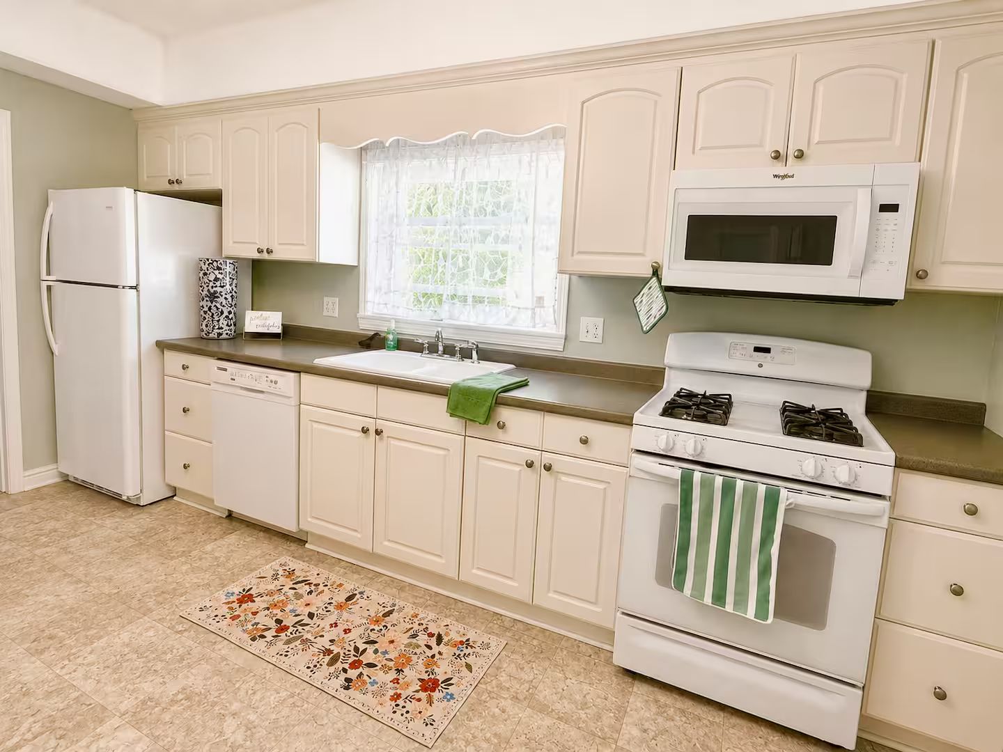 A kitchen with white cabinets, a refrigerator, a stove, a microwave, and a window with a curtain.