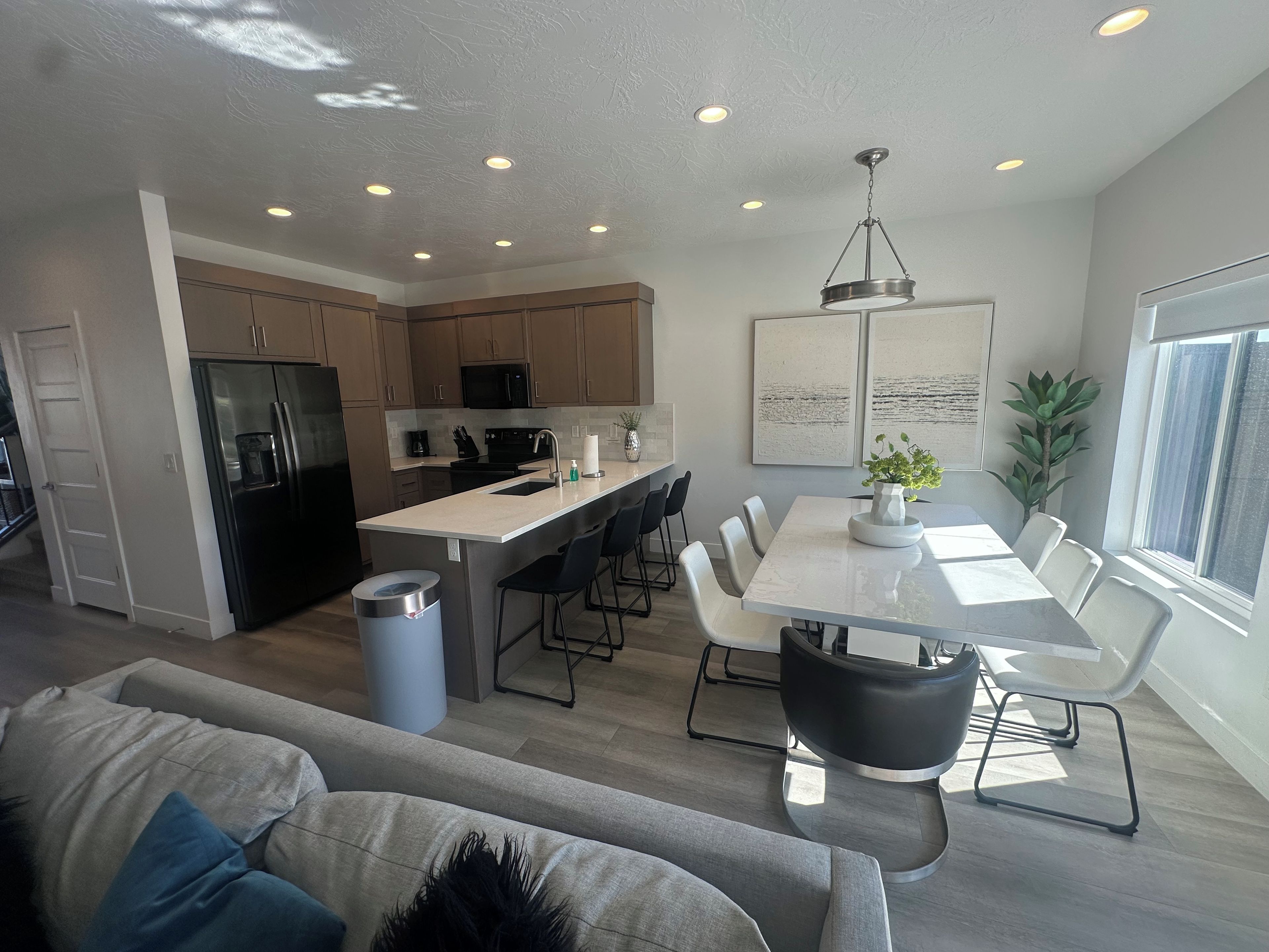 A modern kitchen with a central island, a dining area with a white table and chairs, and a living room with a grey sofa.