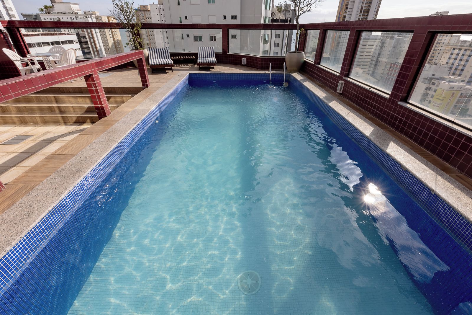 A rooftop swimming pool with a clear blue water surface, surrounded by a red-tiled deck and a glass balustrade.