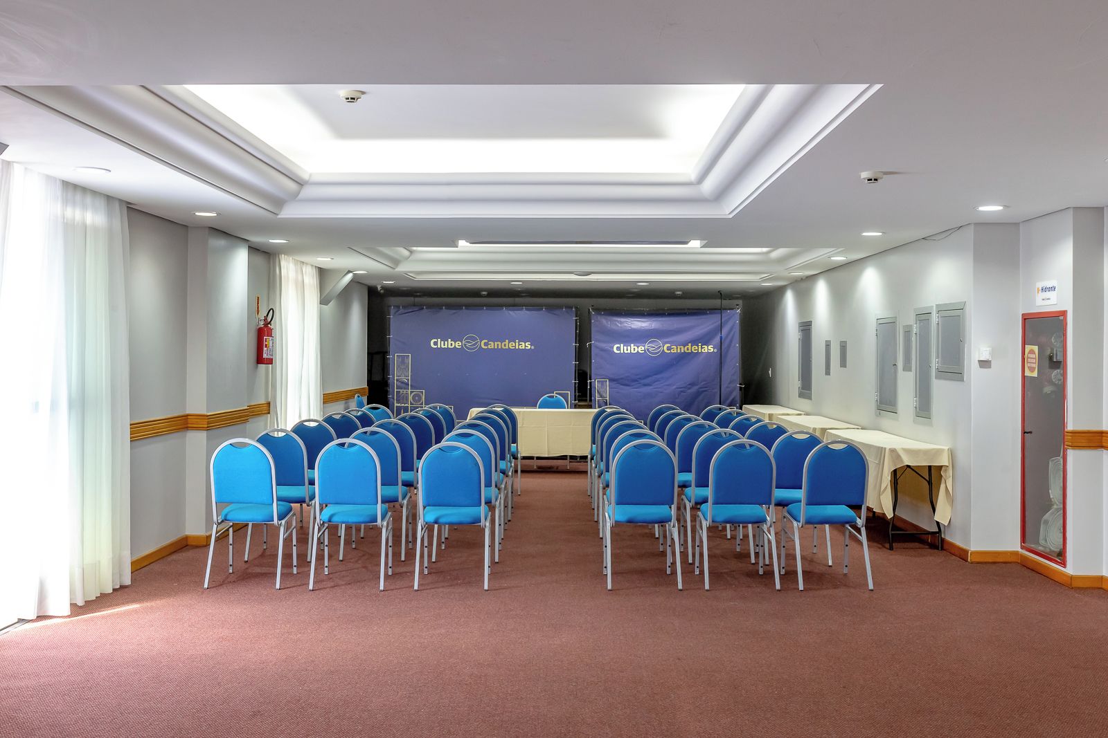 A conference room with rows of blue chairs, a stage with a banner, and a fire extinguisher on the wall.