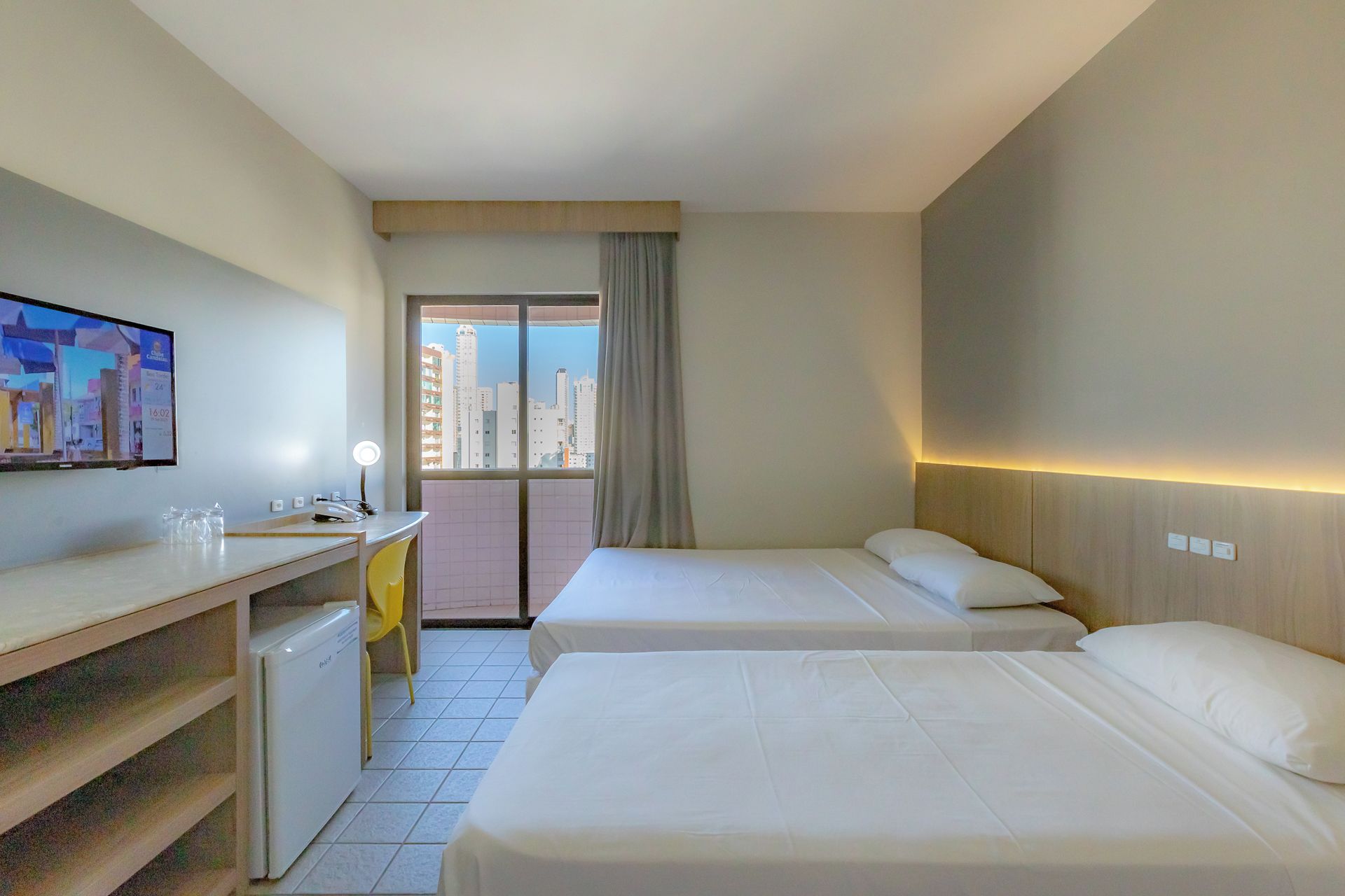 A hotel room with two beds, a desk, a television, and a view of the city.