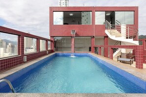 Una piscina con solárium de baldosas rojas, una escalera y vistas a la ciudad.