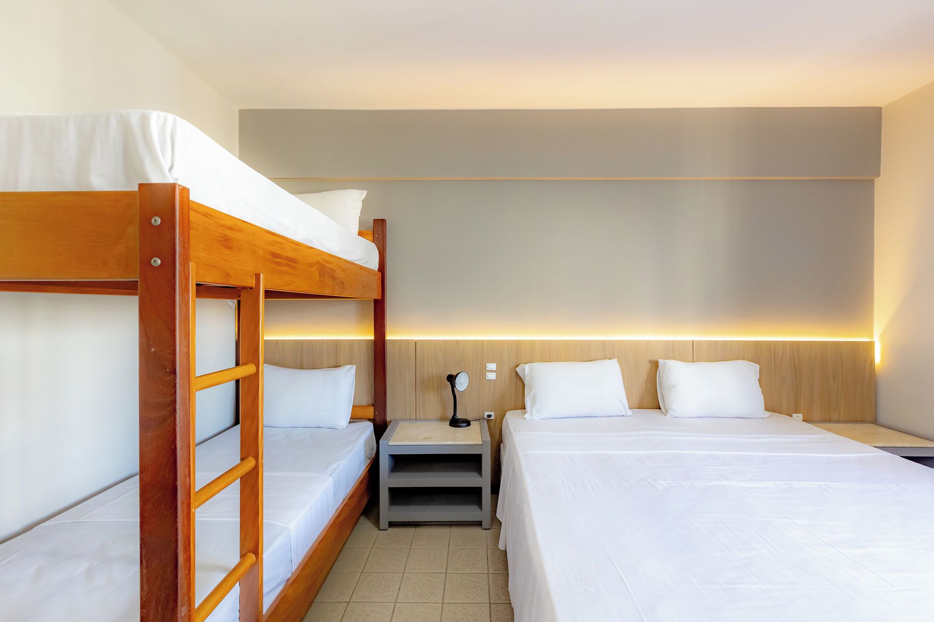A hotel room with a bunk bed, a single bed, a bedside table with a lamp, and a wall-mounted light.